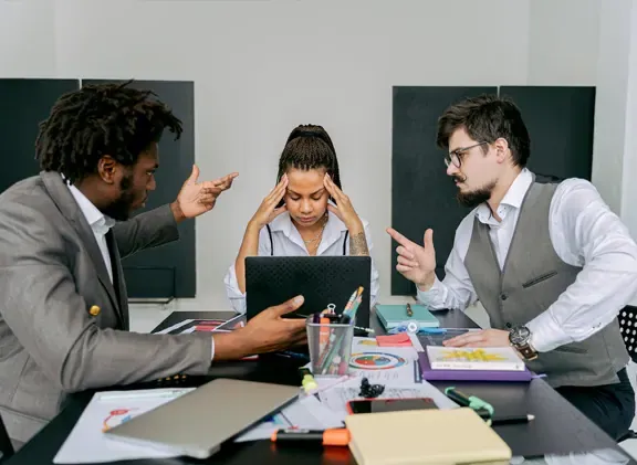 Stressed woman holds her head at a desk while two male colleagues argue and gesture over her laptop