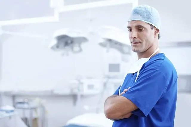 Male surgeon in blue scrubs and cap standing with arms crossed in an operating room with overhead lights