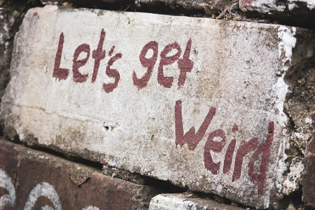 Weathered stone block with red painted handwritten text reading Let's get weird against a brick wall