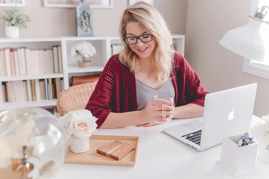 Smiling blonde woman in glasses and red cardigan holding a mug at a white desk with a laptop in a bright home office