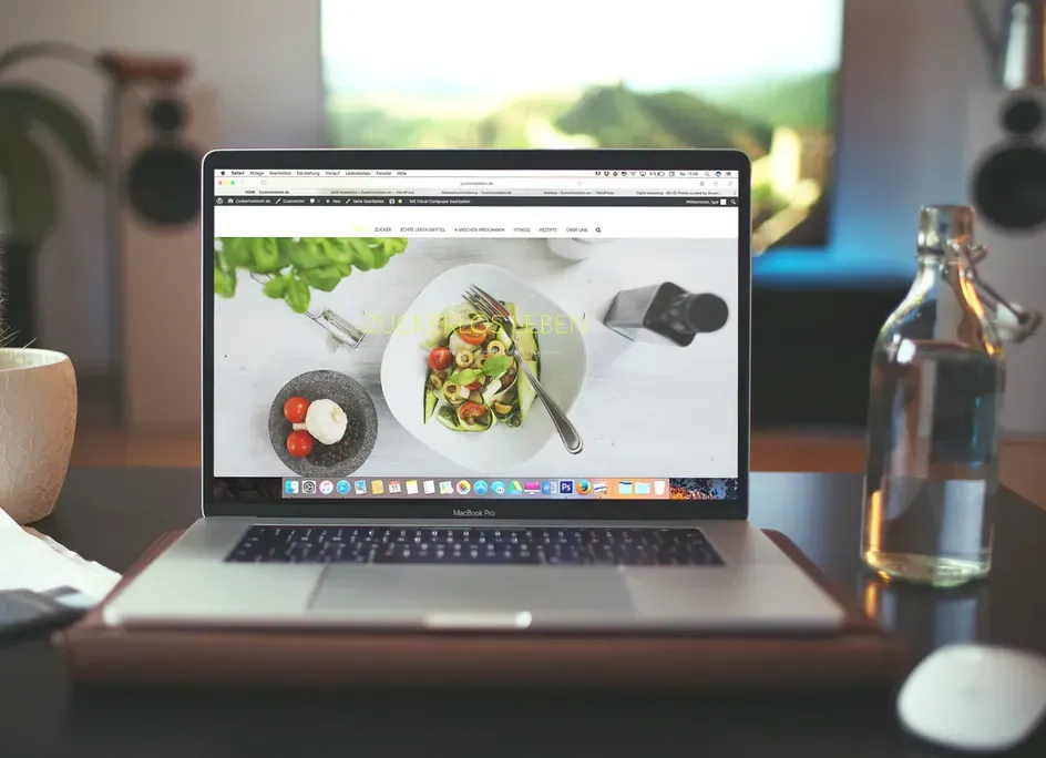 MacBook on a wooden desk displaying a food website with a salad photo, beside a glass water bottle and plant