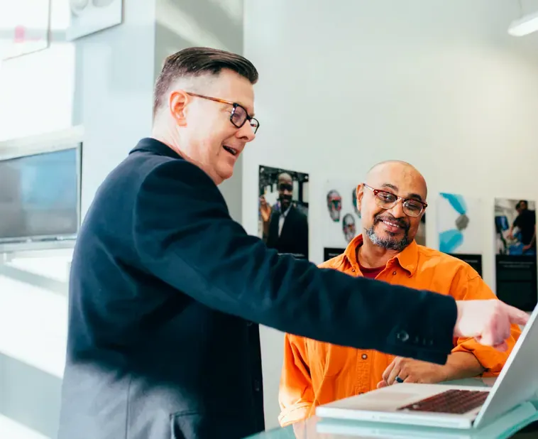Two smiling men in glasses look at a laptop screen together in a bright office with wall posters