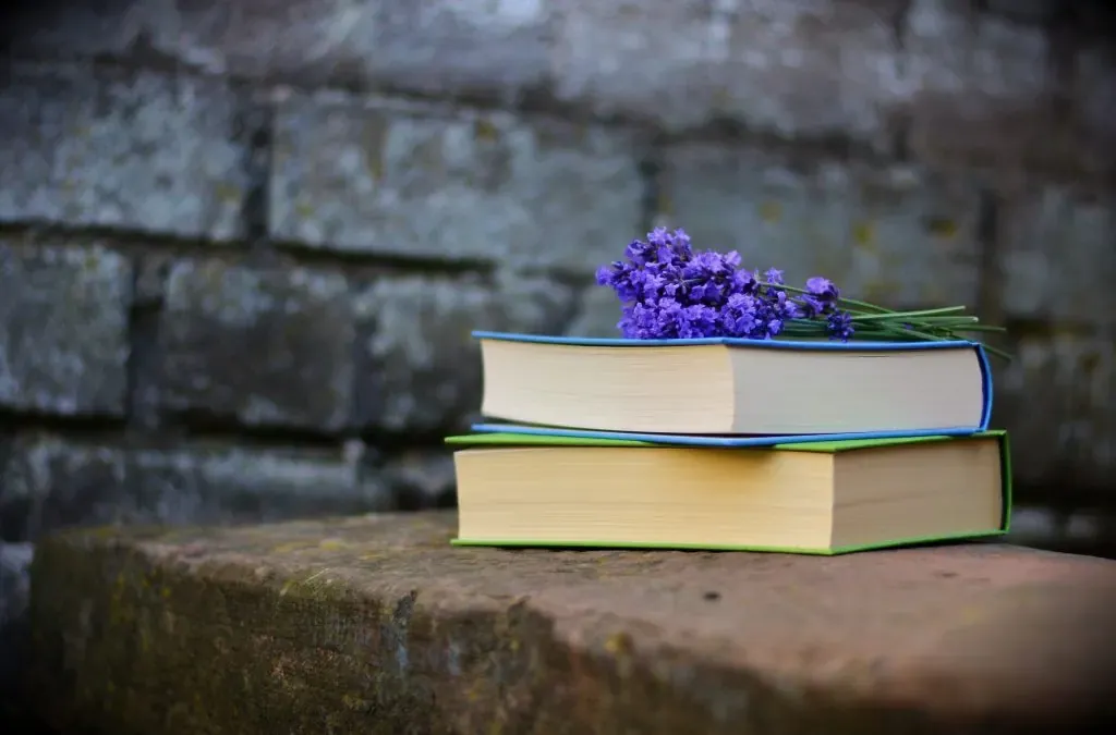Two closed hardcover books stacked on a stone ledge with a sprig of lavender resting on top against a gray brick wall