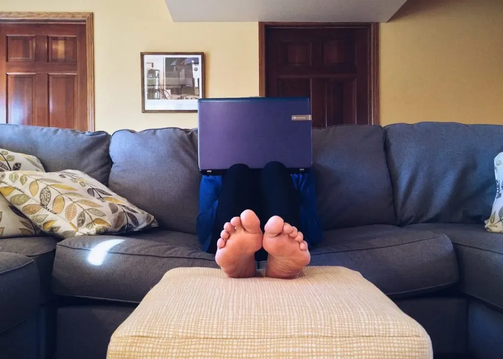 Person reclining on a gray sectional couch with bare feet on an ottoman, laptop hiding their face