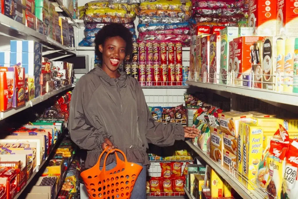 Smiling woman holding an orange shopping basket in a grocery store aisle stocked with cereal and snacks