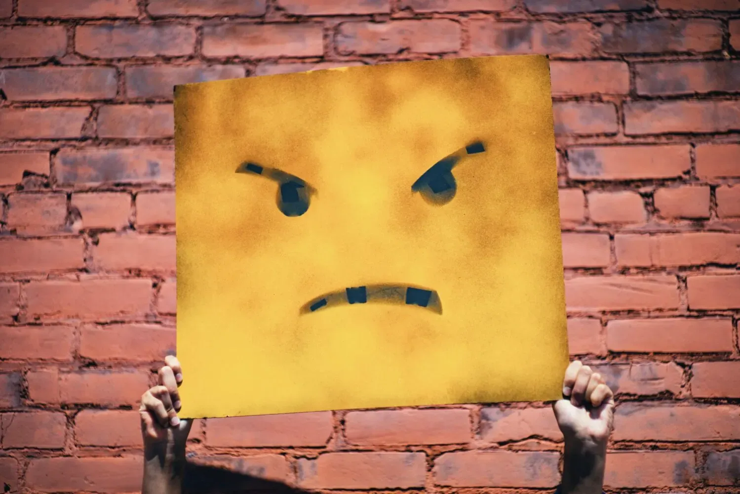 Hands holding up a large yellow sign painted with an angry frowning face against a red brick wall