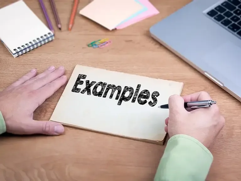 Hands holding a pen over a notepad with the word Examples written on it, beside a laptop and pencils on a wooden desk
