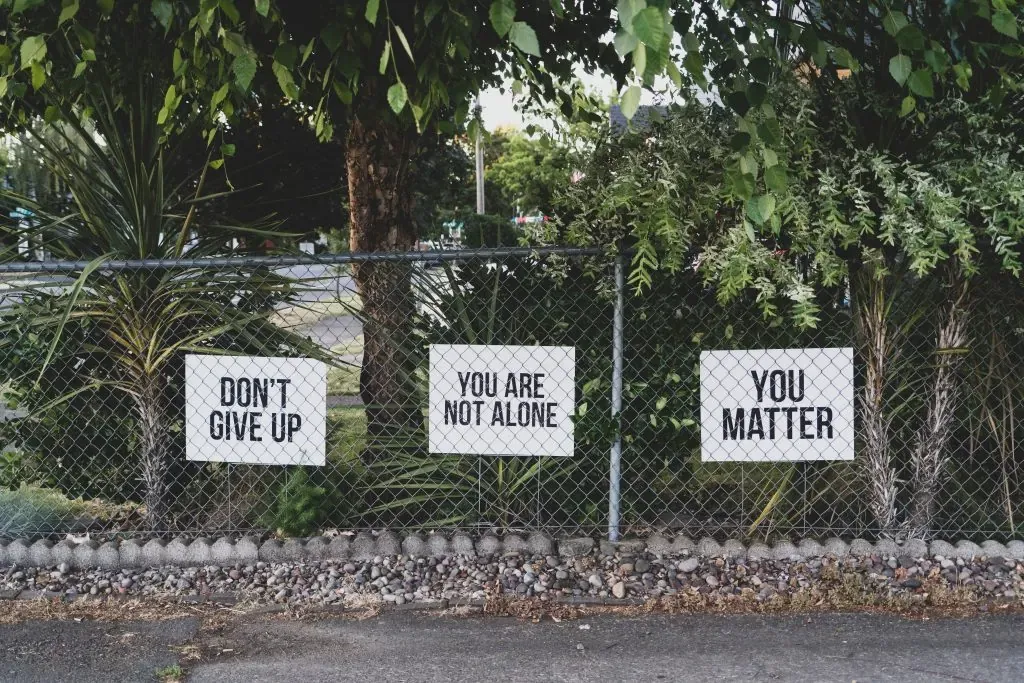 Three signs on a chain link fence reading Don't Give Up, You Are Not Alone, and You Matter among greenery
