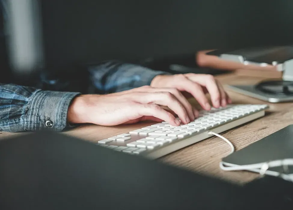 Close-up of a person in a denim shirt typing on a white keyboard at a wooden desk
