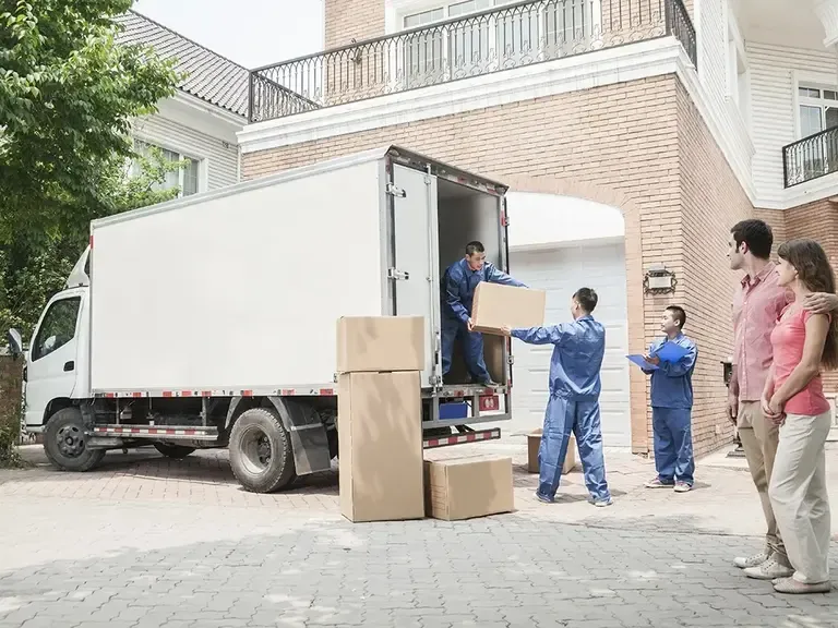 Movers in blue uniforms unload boxes from a white truck while a couple watches outside a brick home