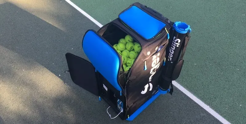 Blue and black Slinger Bag tennis ball launcher filled with yellow balls on a green court surface