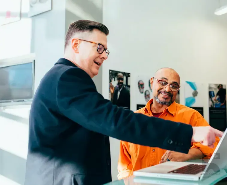 Two smiling men in glasses look at a laptop screen together in a bright office with wall posters