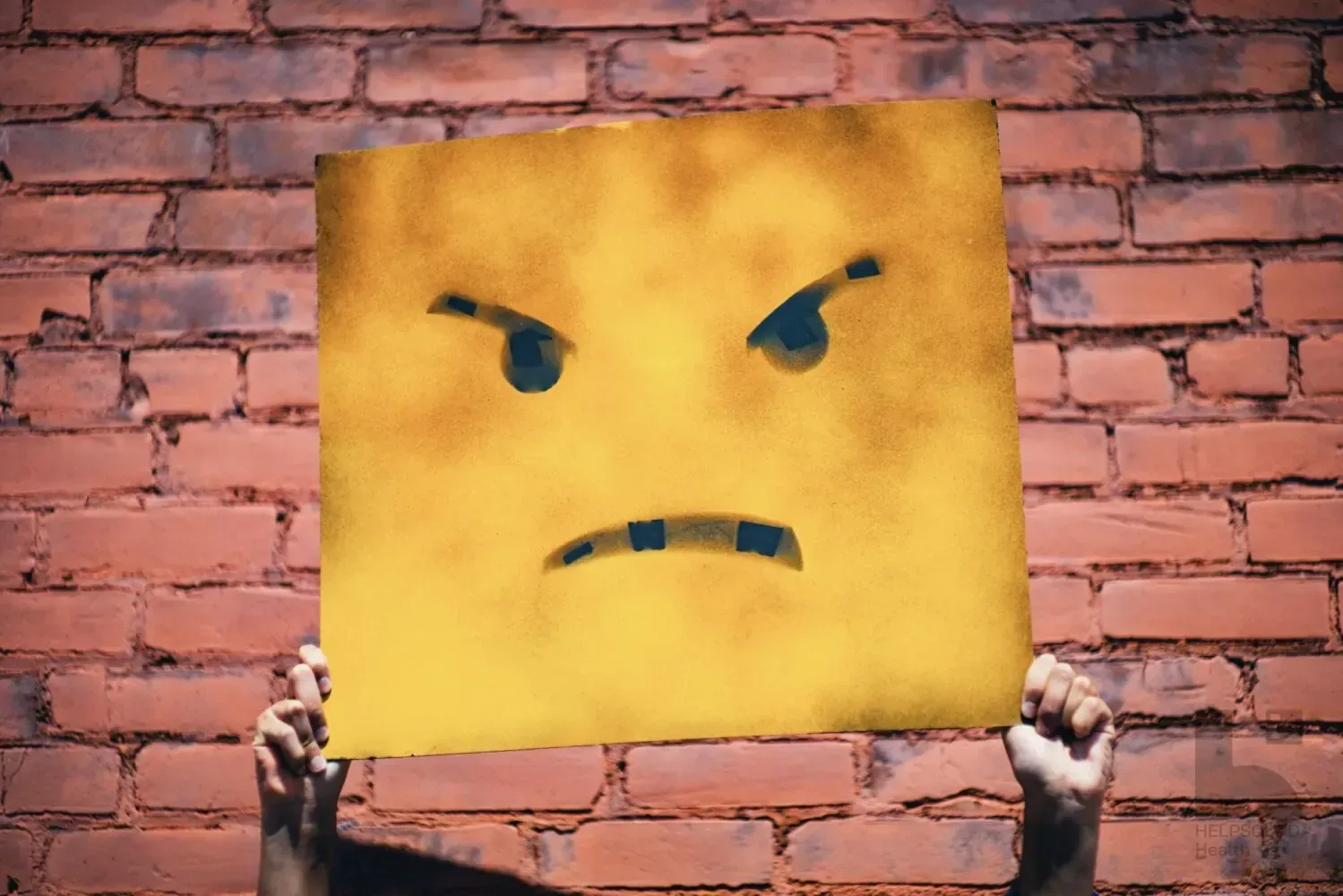 Hands holding up a large yellow sign painted with an angry frowning face against a red brick wall