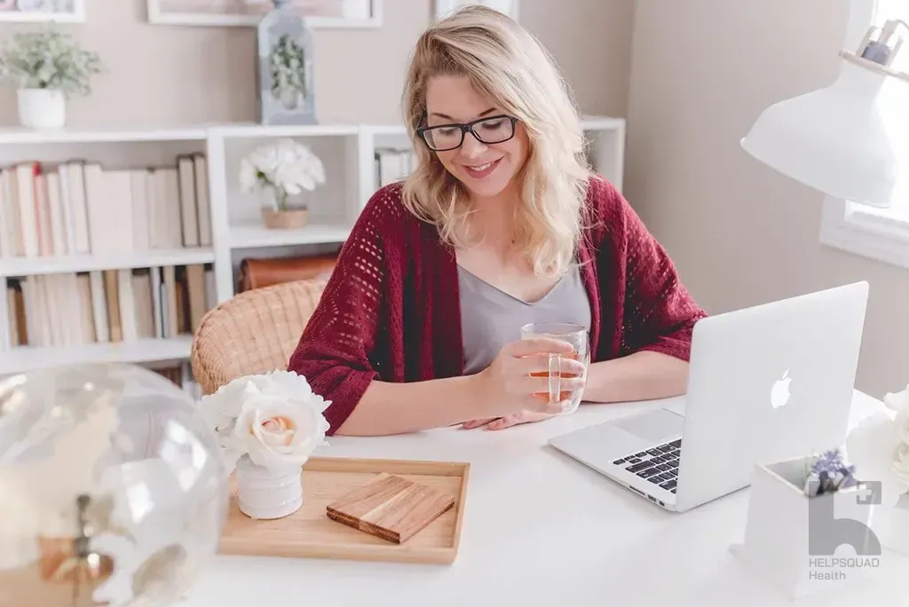 Smiling blonde woman in glasses and red cardigan holding a mug at a white desk with a laptop in a bright home office