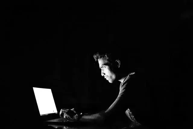 Black and white shot of a man hunched over a glowing laptop screen in a darkened room