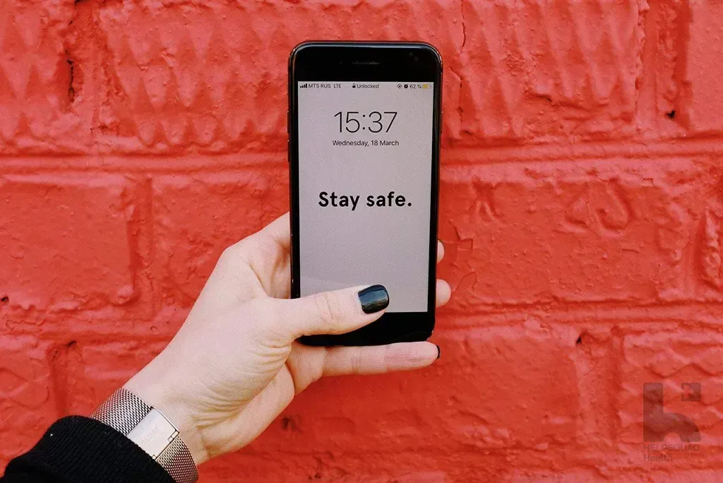 Hand holding a smartphone displaying 15:37 and the message Stay safe against a red painted brick wall