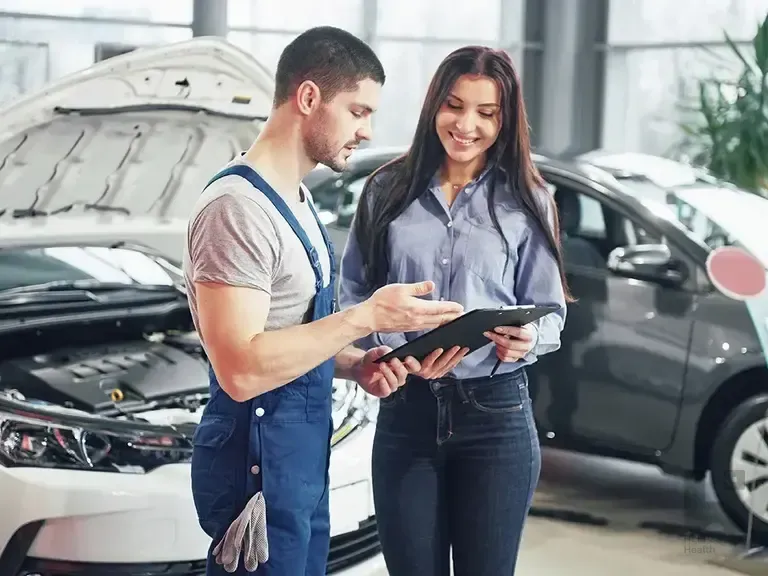 Mechanic in overalls and a smiling woman review a tablet together in front of cars in an auto shop