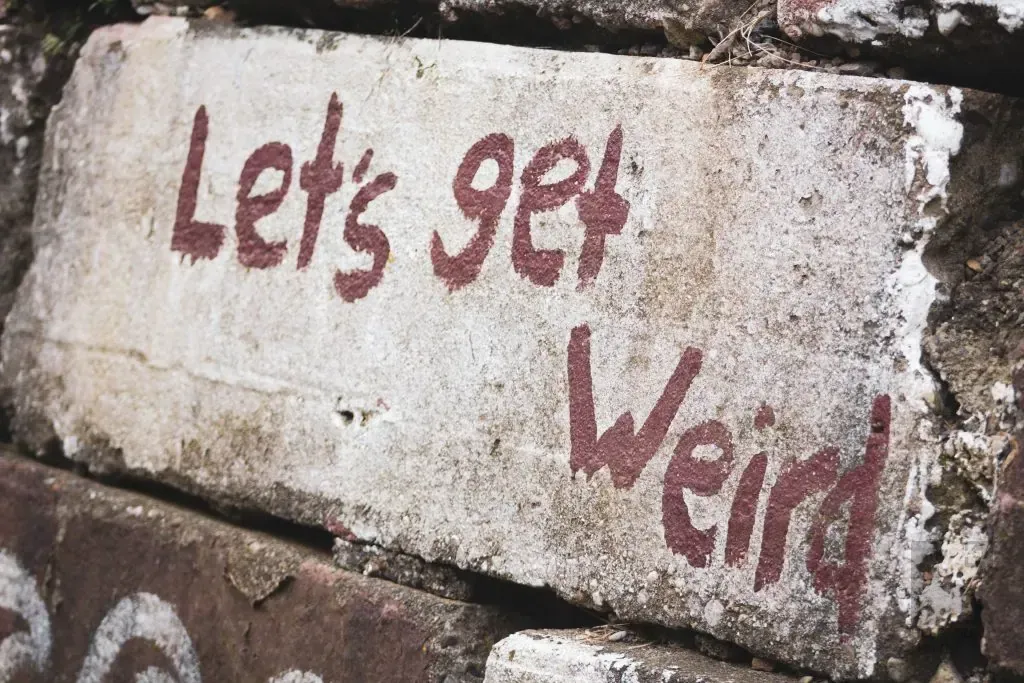 Weathered stone block with red painted handwritten text reading Let's get weird against a brick wall