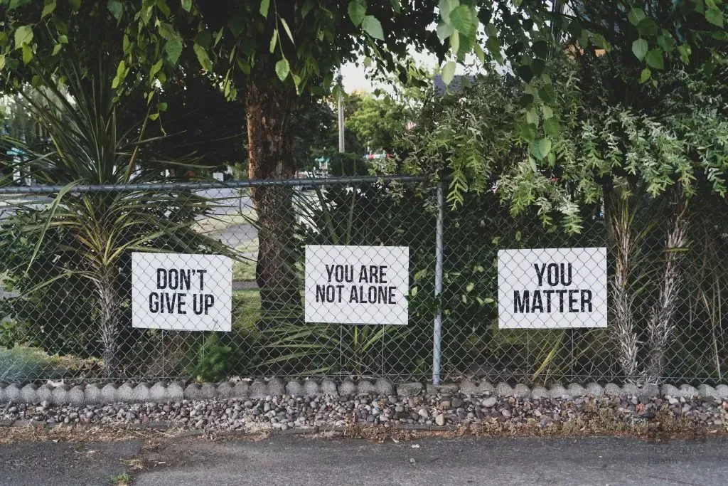 Three signs on a chain link fence reading Don't Give Up, You Are Not Alone, and You Matter among greenery