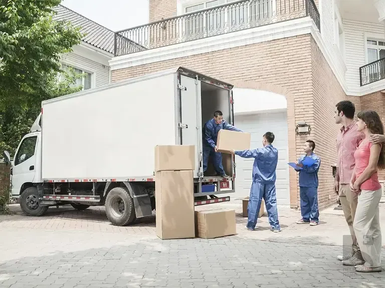 Movers in blue uniforms unload boxes from a white truck while a couple watches outside a brick home