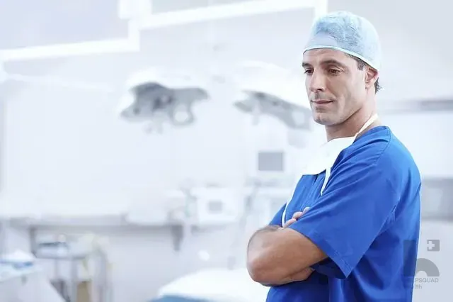 Male surgeon in blue scrubs and cap standing with arms crossed in an operating room with overhead lights