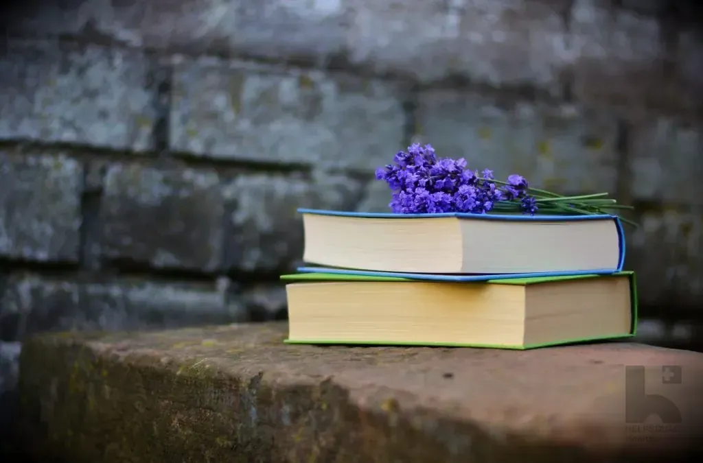 Two closed hardcover books stacked on a stone ledge with a sprig of lavender resting on top against a gray brick wall