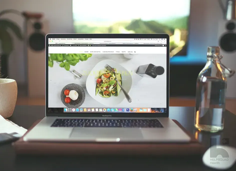 MacBook on a wooden desk displaying a food website with a salad photo, beside a glass water bottle and plant