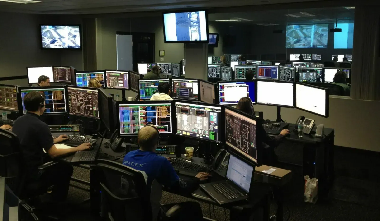 Operators in a dimly lit control room monitoring banks of computer screens and overhead displays around the clock