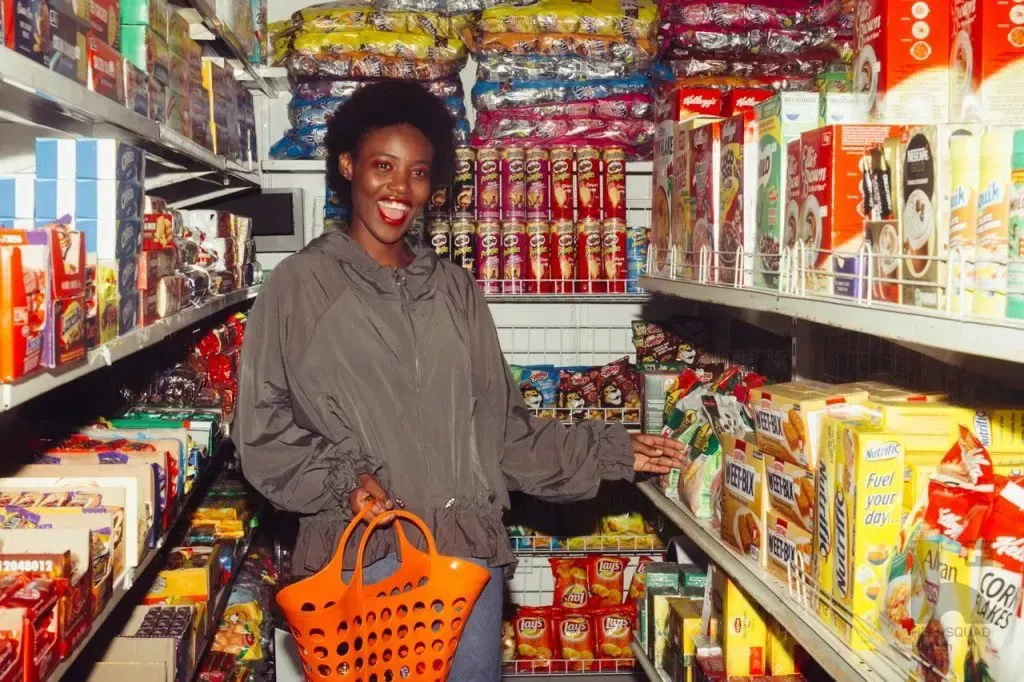 Smiling woman holding an orange shopping basket in a grocery store aisle stocked with cereal and snacks