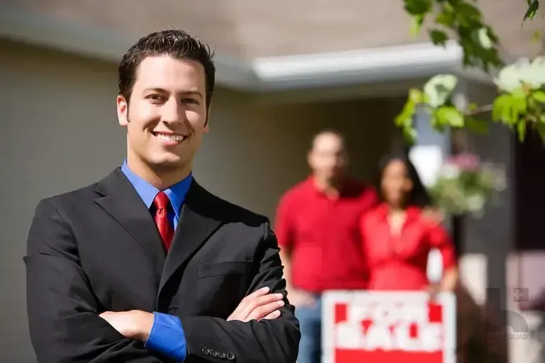 Smiling real estate agent in a suit with arms crossed, a couple and For Sale sign behind him at a house