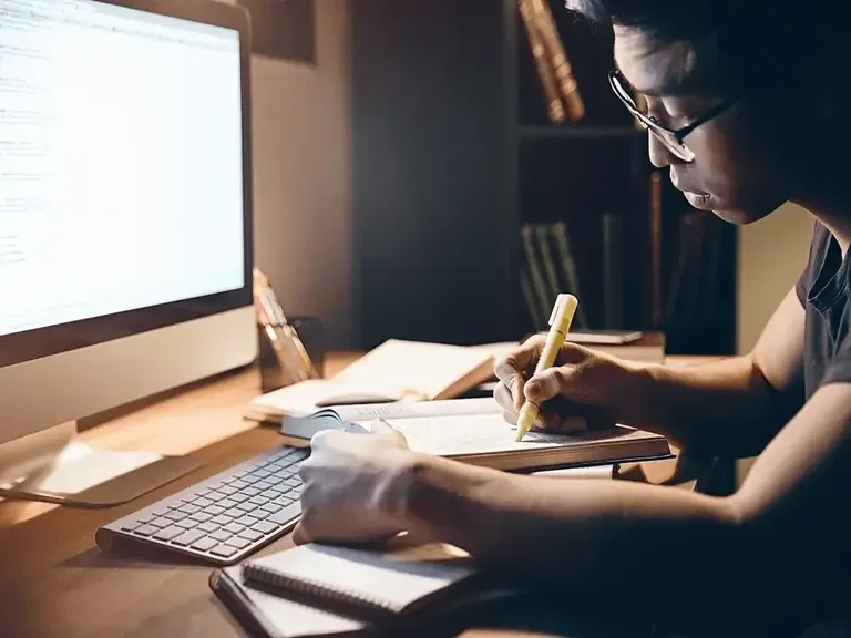 Young man in glasses writing notes in a notebook at a desk beside a large desktop monitor and keyboard