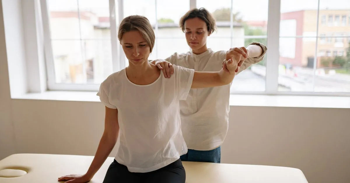 A therapist supports a woman's outstretched arm as she sits on a treatment table.