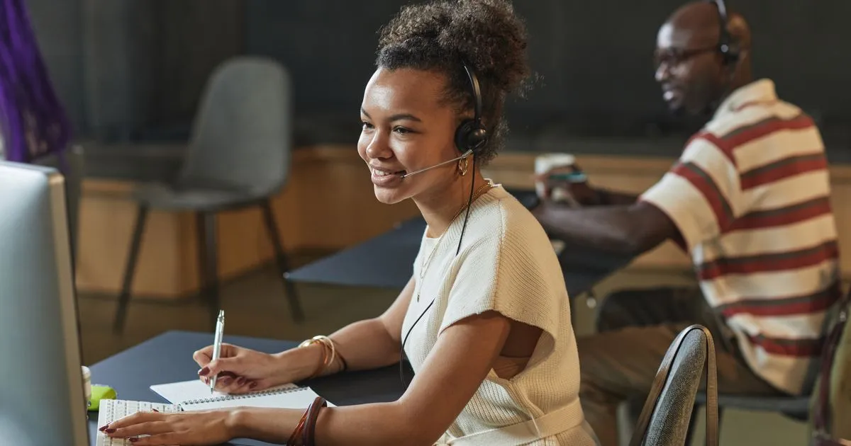 Smiling woman wearing a headset takes notes at a computer while a colleague works nearby.