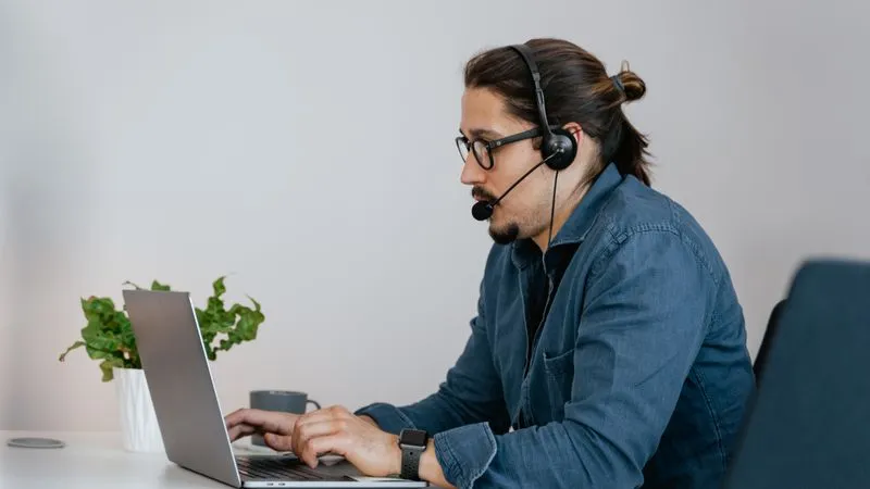 Man with glasses wearing a headset and typing on a laptop at a white desk.
