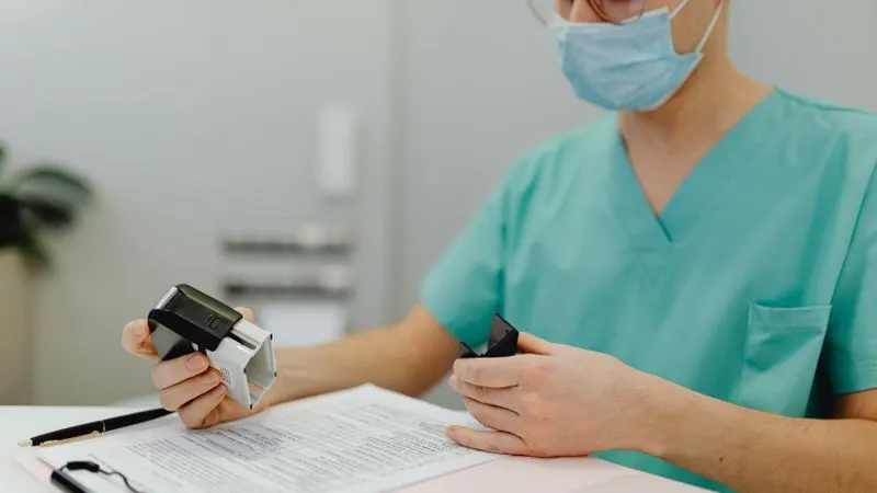 Healthcare worker in teal scrubs and a mask holding a stamp over paperwork on a clipboard.