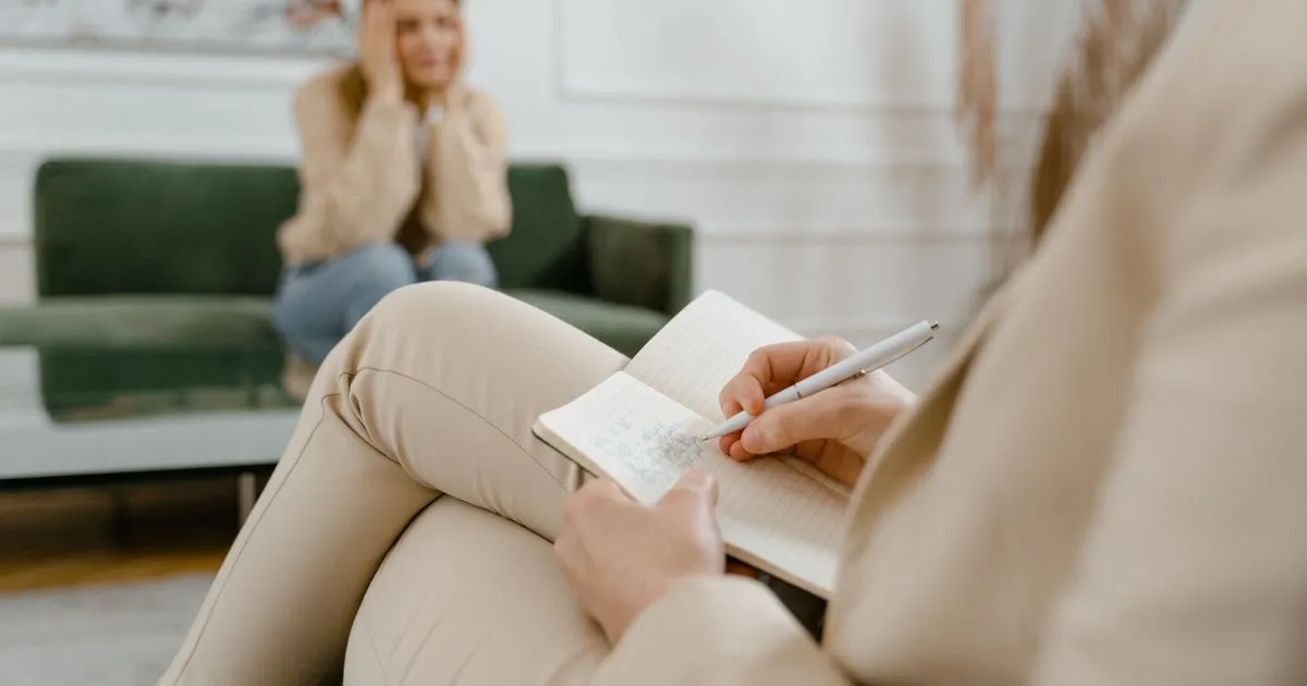 A therapist writes notes in a notebook while a client sits on a green sofa.